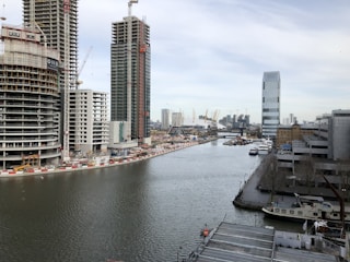 A river or canal runs through a cityscape with construction activity. Tall buildings and cranes dominate the background, with some structures under construction. A skyscraper stands prominently on the right, while smaller industrial buildings and several moored boats line the waterway.