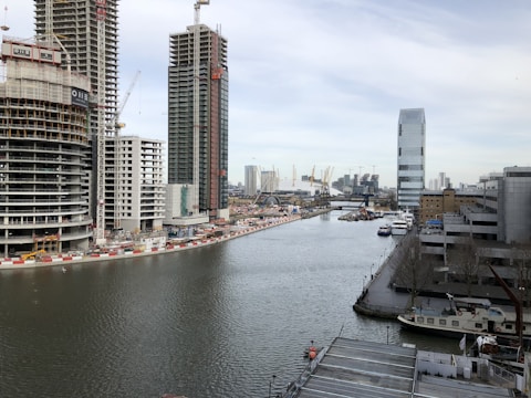 A river or canal runs through a cityscape with construction activity. Tall buildings and cranes dominate the background, with some structures under construction. A skyscraper stands prominently on the right, while smaller industrial buildings and several moored boats line the waterway.