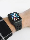 A smartwatch with a black strap is displayed on a wrist, featuring a grid of colorful app icons on its screen. The background is plain, and the wrist is partially covered by a plaid shirt sleeve.