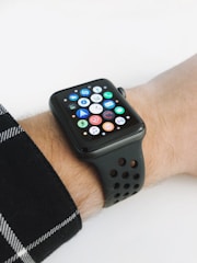 A smartwatch with a black strap is displayed on a wrist, featuring a grid of colorful app icons on its screen. The background is plain, and the wrist is partially covered by a plaid shirt sleeve.