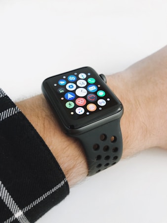 A smartwatch with a black strap is displayed on a wrist, featuring a grid of colorful app icons on its screen. The background is plain, and the wrist is partially covered by a plaid shirt sleeve.