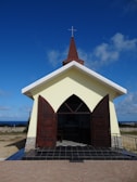 A small chapel with a beige facade and a dark brown pointed roof, topped with a cross. The chapel has open wooden double doors and sits against a backdrop of a clear blue sky and a landscape featuring the ocean in the distance.