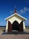A small chapel with a beige facade and a dark brown pointed roof, topped with a cross. The chapel has open wooden double doors and sits against a backdrop of a clear blue sky and a landscape featuring the ocean in the distance.
