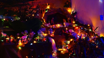 A detailed nativity scene with various figurines of animals and people. String lights of various colors illuminate the scene, creating a warm and festive atmosphere. Small houses and other structures are also present, enhancing the setting. The background appears to be a painted mural, adding depth to the scene.