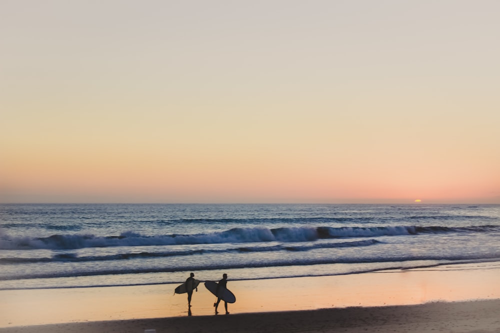 two person carrying surfboards while walking on beach shore