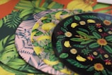 A collection of vibrant, patterned plates with tropical designs including leaves, bananas, and flowers. The plates are stacked in a layered manner, each having distinct colors and patterns featuring lush green foliage and detailed floral motifs.