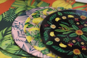 A collection of vibrant, patterned plates with tropical designs including leaves, bananas, and flowers. The plates are stacked in a layered manner, each having distinct colors and patterns featuring lush green foliage and detailed floral motifs.