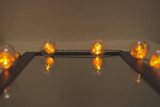 A row of glowing filament bulbs arranged along the edge of a reflective surface. The bulbs emit a warm orange light, and their reflections create a symmetrical effect.