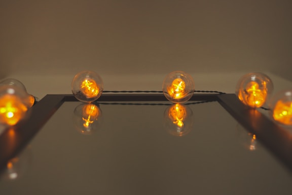 A row of glowing filament bulbs arranged along the edge of a reflective surface. The bulbs emit a warm orange light, and their reflections create a symmetrical effect.
