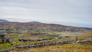 A sweeping view of the lush Irish countryside under soft morning light, with rolling green hills and a distant stone wall.