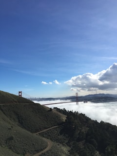 A view of the Golden Gate Bridge spanning across a bay with low-lying fog partially obscuring the structure. The bridge is flanked by hills covered in green vegetation, and the sky above is bright blue with scattered clouds.