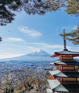 A traditional Japanese pagoda stands prominently in the foreground, with intricate architecture and red accents. In the background, the iconic snow-capped Mount Fuji rises majestically under a clear blue sky. Below, a sprawling cityscape stretches out, framed by lush green trees, some displaying autumn foliage.