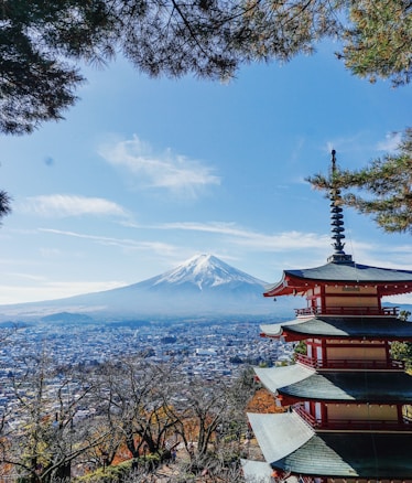 A traditional Japanese pagoda stands prominently in the foreground, with intricate architecture and red accents. In the background, the iconic snow-capped Mount Fuji rises majestically under a clear blue sky. Below, a sprawling cityscape stretches out, framed by lush green trees, some displaying autumn foliage.