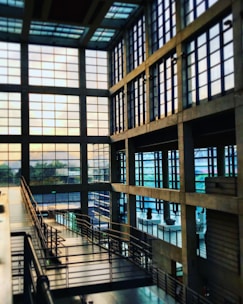 A spacious, modern interior with large grid-like windows allowing natural light to fill the area. The architecture features an industrial style with exposed beams and metallic railings. A walkway or ramp runs diagonally through the space, surrounded by glass and concrete.