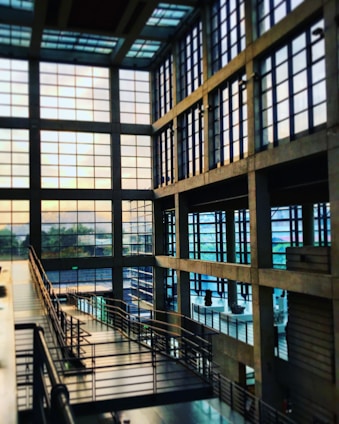 A spacious, modern interior with large grid-like windows allowing natural light to fill the area. The architecture features an industrial style with exposed beams and metallic railings. A walkway or ramp runs diagonally through the space, surrounded by glass and concrete.