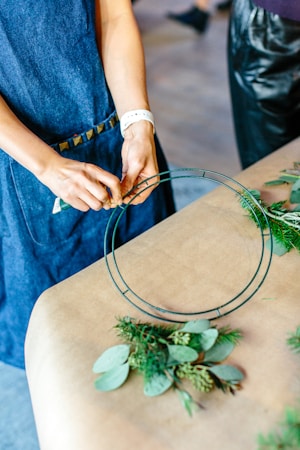 A person wearing a blue denim apron is crafting a wreath. They are holding a green wire frame on a wooden surface with decorative foliage laid out. Some of the plant materials include various green leaves and small branches.