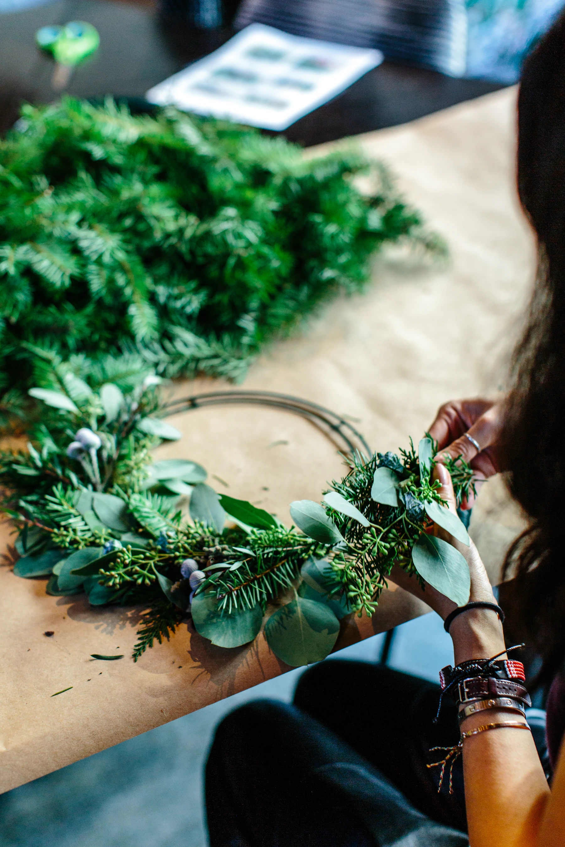 Hands wiring a bundle of fir and eucalyptus onto a wire ring on a paper-covered table