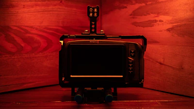 A digital camera setup is displayed against a warm, reddish-brown wooden backdrop. The camera appears to be mounted on a rig with multiple components, including a monitor and control buttons that are visible on the right side. The lighting casts a soft red glow over the entire scene, enhancing the wood grain texture.