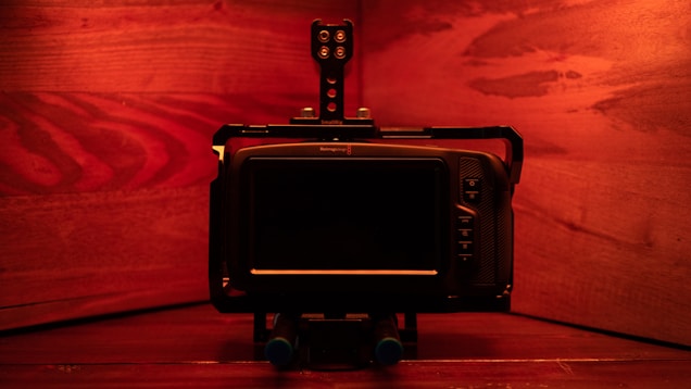 A digital camera setup is displayed against a warm, reddish-brown wooden backdrop. The camera appears to be mounted on a rig with multiple components, including a monitor and control buttons that are visible on the right side. The lighting casts a soft red glow over the entire scene, enhancing the wood grain texture.