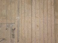 Aerial view of expansive agricultural fields with parallel lines indicating rows of planting or harvesting. Three small figures, possibly workers, are dispersed across the field wearing distinctive clothing that contrasts with the earthy tones of the ground. The fields have a patchy appearance, possibly indicating different stages of crop growth or soil moisture.