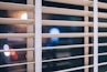 Wooden blinds partially cover a window at night, through which blurred lights from outside are visible.