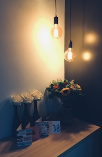 A warmly lit scene with two hanging light bulbs casting a soft glow. On a wooden surface, there are three tall vases each holding a white rose. Beside them is a collection of greeting cards, one of which says 'Happy Birthday', along with a card displaying the number '38'. To the right, a glass vase is filled with a colorful bouquet, including sunflowers and various greenery.