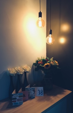 A warmly lit scene with two hanging light bulbs casting a soft glow. On a wooden surface, there are three tall vases each holding a white rose. Beside them is a collection of greeting cards, one of which says 'Happy Birthday', along with a card displaying the number '38'. To the right, a glass vase is filled with a colorful bouquet, including sunflowers and various greenery.