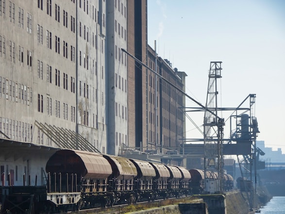 A series of industrial train cars are parked alongside a large, multi-story warehouse building. The building has numerous windows and a slightly worn facade. Overhead cranes and industrial equipment are also visible, suggesting a setting in a port or industrial area.