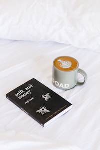 A black book titled 'Milk and Honey' by Rupi Kaur is resting on a white bedspread next to a green mug of coffee with latte art on top. The latte art features a leaf pattern, and the mug has the word 'ROAD' printed on it.