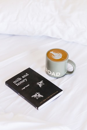 A black book titled 'Milk and Honey' by Rupi Kaur is resting on a white bedspread next to a green mug of coffee with latte art on top. The latte art features a leaf pattern, and the mug has the word 'ROAD' printed on it.