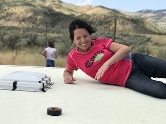 A woman wearing a bright pink sports shirt is smiling and reclining on a light-colored surface. A roll of black tape is near her hand. In the background, a person stands facing away, with a wide-spanning landscape of hills covered in greenery.