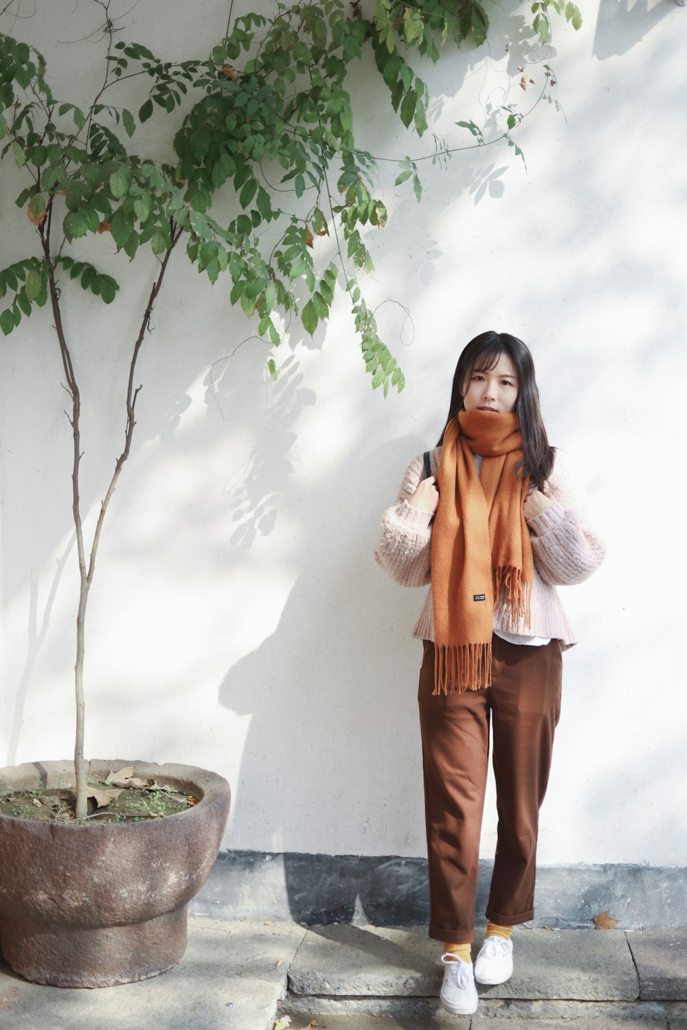 woman with orange scarf stands beside potted tree