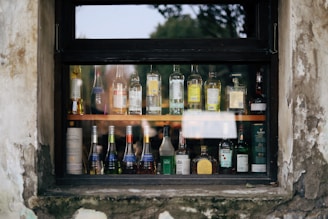 A variety of liquor bottles are displayed on shelves behind a window. The window frame is weathered, with signs of peeling paint and age. The bottles are of different brands and sizes, arranged neatly in rows, showcasing their labels and contents.