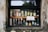 A variety of liquor bottles are displayed on shelves behind a window. The window frame is weathered, with signs of peeling paint and age. The bottles are of different brands and sizes, arranged neatly in rows, showcasing their labels and contents.