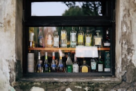 A variety of liquor bottles are displayed on shelves behind a window. The window frame is weathered, with signs of peeling paint and age. The bottles are of different brands and sizes, arranged neatly in rows, showcasing their labels and contents.