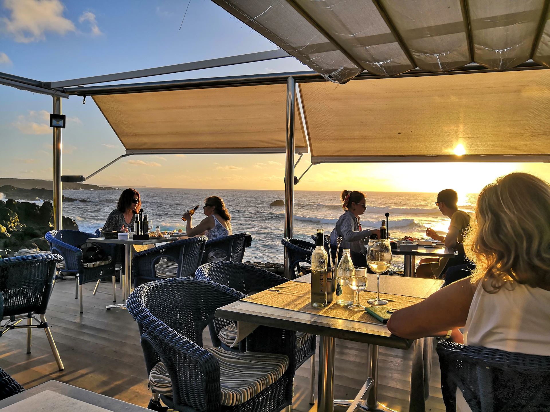 A cozy deck scene at sunset with passengers enjoying drinks and the ocean horizon glowing in warm colors.