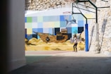 A young girl with colorful toy wings and a backpack stands in a partially enclosed courtyard. The walls are adorned with a vibrant mural featuring an underwater scene with a shipwreck, fish, seaweed, and a mosaic of squares in various pastel shades. A basketball hoop is attached to one wall, with support beams around it.