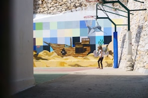 A young girl with colorful toy wings and a backpack stands in a partially enclosed courtyard. The walls are adorned with a vibrant mural featuring an underwater scene with a shipwreck, fish, seaweed, and a mosaic of squares in various pastel shades. A basketball hoop is attached to one wall, with support beams around it.