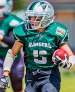 A player in football gear runs with the ball, wearing a green jersey with 'Rangers' written on it and the number 5. The helmet has a distinctive logo. Sunlight highlights the player's movement on the field.