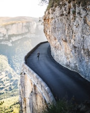 A narrow, winding road hugs the edge of a steep cliff, with a person running on it. The surrounding landscape features rugged rock formations and expansive views of lush green valleys. The scene conveys a sense of adventure and exhilaration, emphasizing the dramatic contrast between the safety of the road and the precipitous drop-off.