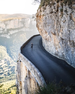 A narrow, winding road hugs the edge of a steep cliff, with a person running on it. The surrounding landscape features rugged rock formations and expansive views of lush green valleys. The scene conveys a sense of adventure and exhilaration, emphasizing the dramatic contrast between the safety of the road and the precipitous drop-off.