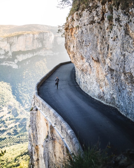 A narrow, winding road hugs the edge of a steep cliff, with a person running on it. The surrounding landscape features rugged rock formations and expansive views of lush green valleys. The scene conveys a sense of adventure and exhilaration, emphasizing the dramatic contrast between the safety of the road and the precipitous drop-off.