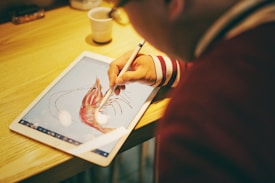 A person is using a stylus to draw a detailed shrimp illustration on a digital tablet placed on a wooden table. The individual's focus and concentration are evident as they work on the digital artwork, capturing intricate details of the shrimp.