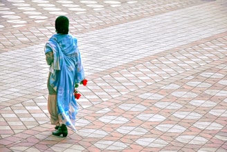 A person wearing a blue shawl and a head covering is walking across a patterned pavement. The person is holding a few red flowers in their hand. The pavement has a symmetrical design with alternating light and dark tiles in diamond shapes.