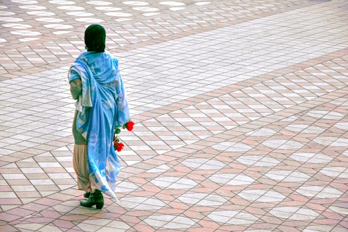 A person wearing a blue shawl and a head covering is walking across a patterned pavement. The person is holding a few red flowers in their hand. The pavement has a symmetrical design with alternating light and dark tiles in diamond shapes.