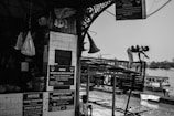 A black-and-white scene captures a busy riverside construction site. A worker stands on a metal structure with a tool in hand, engaged in repairs. Notices and announcements are posted on a tiled wall of a nearby market area, next to hanging bags and baskets. Boats are moored along the riverbank in the background.