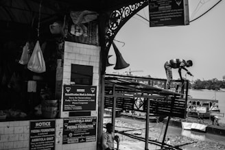 A black-and-white scene captures a busy riverside construction site. A worker stands on a metal structure with a tool in hand, engaged in repairs. Notices and announcements are posted on a tiled wall of a nearby market area, next to hanging bags and baskets. Boats are moored along the riverbank in the background.