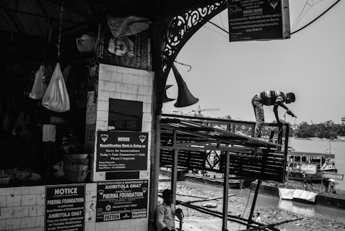 A black-and-white scene captures a busy riverside construction site. A worker stands on a metal structure with a tool in hand, engaged in repairs. Notices and announcements are posted on a tiled wall of a nearby market area, next to hanging bags and baskets. Boats are moored along the riverbank in the background.