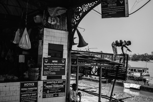 A black-and-white scene captures a busy riverside construction site. A worker stands on a metal structure with a tool in hand, engaged in repairs. Notices and announcements are posted on a tiled wall of a nearby market area, next to hanging bags and baskets. Boats are moored along the riverbank in the background.