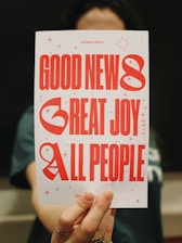 A person is holding a card with the text 'GOOD NEWS, GREAT JOY, ALL PEOPLE' in large red letters. The card is decorated with small red stars and a few lines of smaller text.